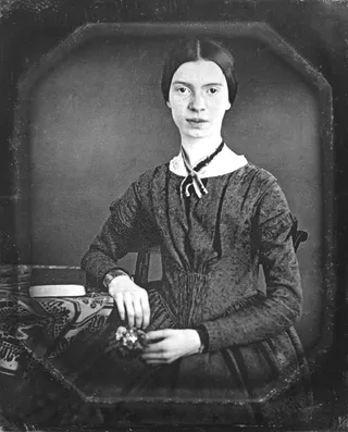 Emily Dickinson seated, holding flowers, with a book on the table beside her, in a nineteenth-century daguerreotype.